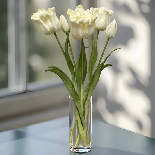 White tulips in a clear glass vase on a reflective surface with a blurred window background