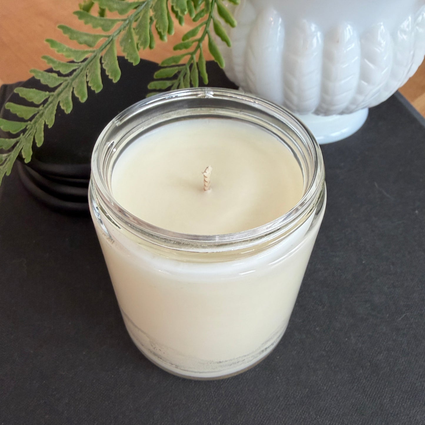White candle in a glass jar with a white decorative object and greenery on a dark surface